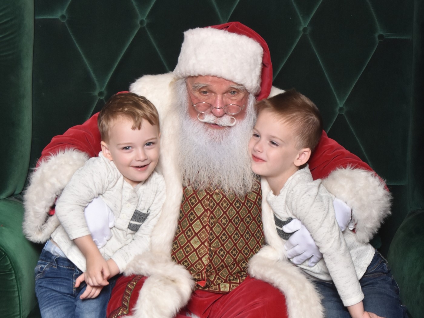 Santa Claus in a festive red and white outfit sits on a green velvet chair, flanked by two smiling young boys.